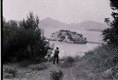Two figures in winter attire stand on a grassy slope overlooking a rocky island in a frozen lake, framed by snow-laden trees....