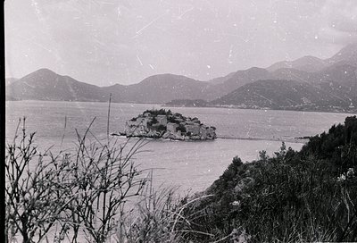Vintage black-and-white coastal scene featuring a small island fortress surrounded by calm waters, framed by rugged hills. Ar...