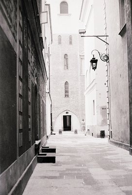 Narrow medieval alleyway with stone-paved path, flanked by tall, weathered buildings. Gothic-style arched doorway at end, fla...