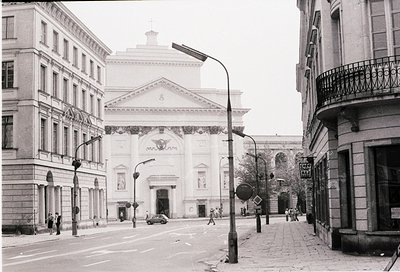 Neoclassical urban plaza featuring grand entrance archway with "МАРКС" signage, flanked by symmetrical stone buildings. Pedes...