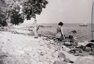 Black-and-white seaside scene featuring two individuals on rocky shore, mid-20th century. Woman in sleeveless dress and short...