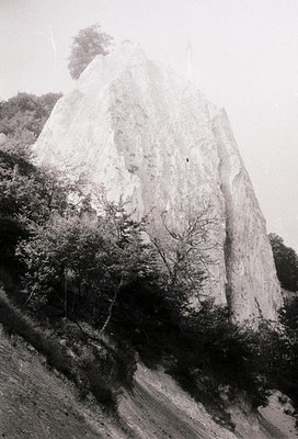 Monolithic rock formation rising sharply from forested terrain, likely limestone. Vegetation includes sparse trees and shrubs...