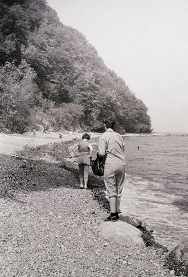 Two figures in mid-20th-century attire (lightweight trousers, short-sleeved shirts) explore a rocky riverside path beside den...