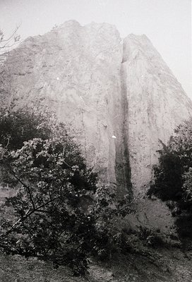 Monolithic limestone cliff with vertical waterfall cascading mid-frame, surrounded by dense foliage. Dramatic natural rock fo...