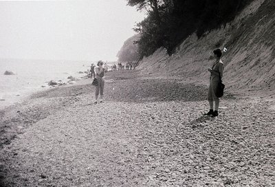 Black-and-white coastal scene showing a gravel path along a rocky cliffside. Two women in mid-20th-century attire (one with a...