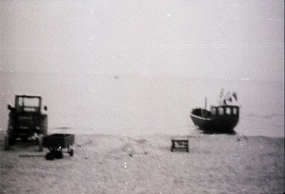 Vintage black-and-white beach scene featuring a small wooden boat, cart, and crate on sandy shore. Minimalist composition sug...
