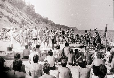 Black-and-white beach scene from mid-20th century, likely 1950s–1960s. Crowded sandy shore with people in swimwear, some seat...