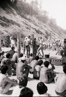 1970s beachside volleyball game with spectators in swimwear, set against rocky cliffs. Crowd seated on sand, players in mid-a...