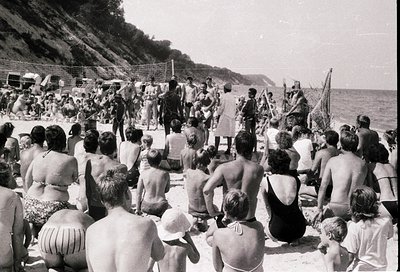 Black-and-white beach scene with a crowd of men, women, and children gathered on sand near a rocky cliffside. Military person...