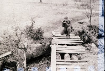 A lone individual sits on a makeshift wooden bridge over a shallow stream in a rural, snow-dusted landscape. The person wears...