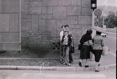 Black-and-white street scene featuring four individuals near a stone monument with Cyrillic text. The monument commemorates a...