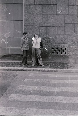 Two young men in 1960s-70s streetwear pose near a Soviet-era concrete wall. One wears a jacket with a pin; Cyrillic text ("КУ...