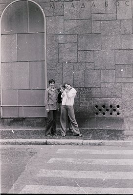 Two men pose against a stone wall with Cyrillic text in 1960s-70s Sofia, Bulgaria. The wall bears Soviet-era inscriptions. On...
