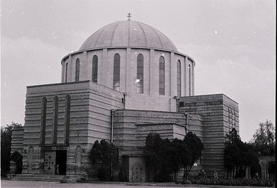 Mid-century modernist church with cylindrical drum dome and brutalist concrete façade. Cross-topped central dome flanked by r...