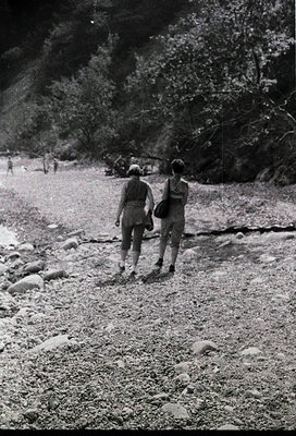 Couple hiking on rocky alpine trail, mid-20th century. Man in light jacket, woman in dark dress. Lush forest backdrop with di...