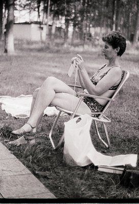Mid-century outdoor scene: Woman in sleeveless dress and sandals knits beside a picnic blanket on grass, seated on a metal ch...