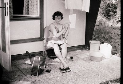 Mid-20th century black-and-white photo of a woman in a bikini writing at a small outdoor table beside a mobile home. Notebook...