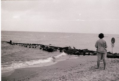 Child stands on a seaside promenade with concrete pier, waves crashing against stone breakwater. Overcast sky suggests mid-20...