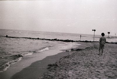 Vintage black-and-white seaside shot of lone figure walking along rocky shore with gentle waves lapping at concrete breakwate...