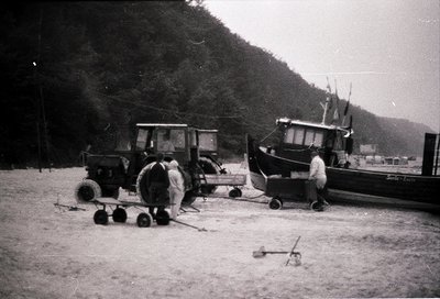 Mid-20th century coastal scene: two men inspecting a vintage tractor and a wooden fishing boat on sandy shore. Hilly, foreste...