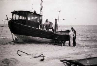 Mid-20th century fishing vessel on a sandy shore, likely Eastern European coastal region. Black hull with enclosed cabin, anc...