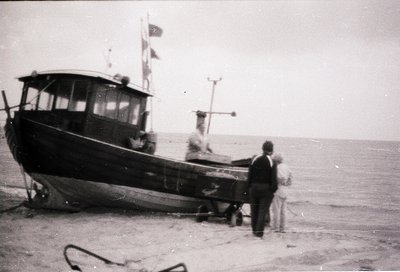 Black-and-white mid-20th century fishing boat on a sandy shore, likely Eastern European coastal region. Two men in dark suits...