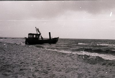 Vintage black-and-white image of a small wooden fishing boat grounded on a sandy shore, waves crashing nearby. Open sea and c...