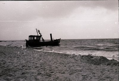 Vintage black-and-white photo of a small wooden fishing vessel grounded on a sandy shore, waves crashing nearby. Mid-20th cen...