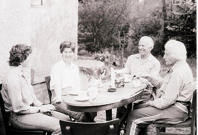 Four adults seated at an outdoor café table, mid-1970s. Lighting suggests daytime, with natural light filtering through trees...