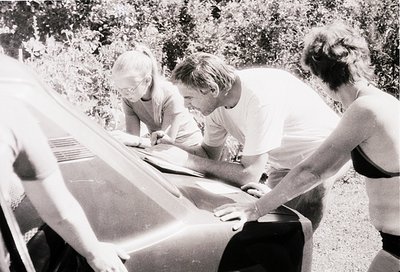 Three individuals engage in outdoor maintenance on a vintage sailboat, likely mid-20th century. The man in the center adjusts...