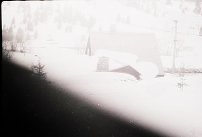 Vintage black-and-white photo of a rural landscape with snow-covered ground and sparse trees. A lone, partially buried struct...