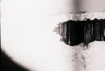 Black-and-white aerial view of a snow-covered cliffside with stacked wooden planks, likely for erosion control or constructio...