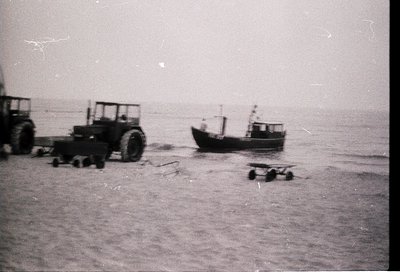 Vintage black-and-white coastal scene featuring a small fishing boat grounded on sandy shore, flanked by industrial tractors ...