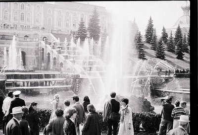 Grand tiered fountain in a grand alpine resort, surrounded by elegant 1950s–60s architecture. Crowds in winter attire—hats, c...