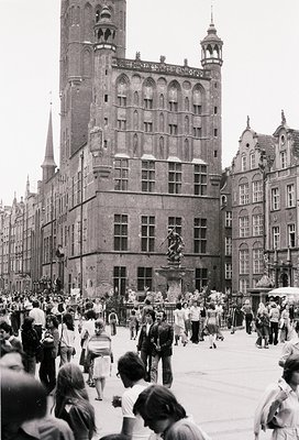 Historic European square bustling with mid-20th century attire. Gothic-style brick buildings with ornate facades and spires d...