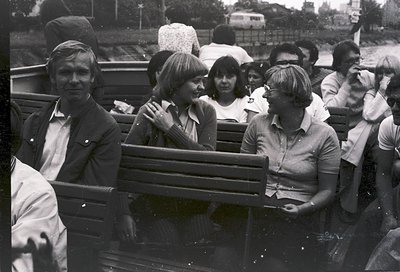 Black-and-white photo of a group seated in an open-air tram or trolley, likely from the **1960s-1970s**. Urban setting with b...