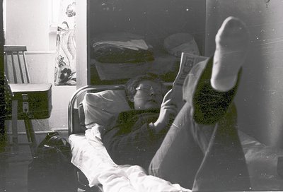 Vintage black-and-white photo of elderly person reclining in hospital bed, reading newspaper. Visible medical equipment (IV s...