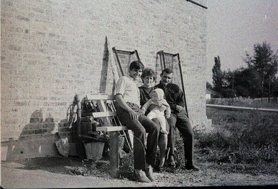Three individuals pose outdoors on a weathered concrete bench beside a brick wall, likely mid-20th century. The man on the le...