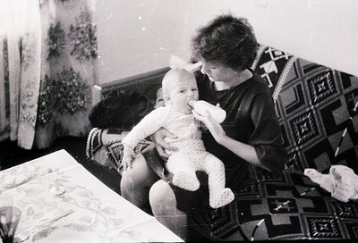 A tender black-and-white moment: an adult feeds a baby seated on a patterned rug, surrounded by vintage floral wallpaper and ...