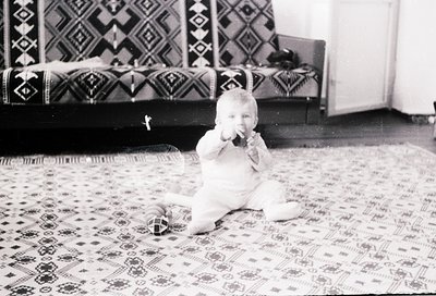 Vintage black-and-white photo of an infant playing on an intricately patterned rug, holding a toy. Geometric textile designs ...