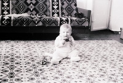 Black-and-white snapshot of an infant in a white knitted outfit, sitting on an intricately patterned rug with geometric desig...