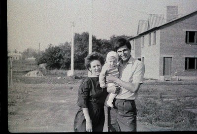 A mid-20th-century black-and-white family portrait: a woman cradles a baby, a man stands beside her in a grassy residential a...