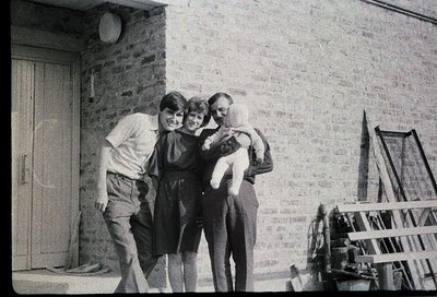 Family portrait outside a brick building, likely residential, with a wooden door and ladder leaning against the wall. Three a...