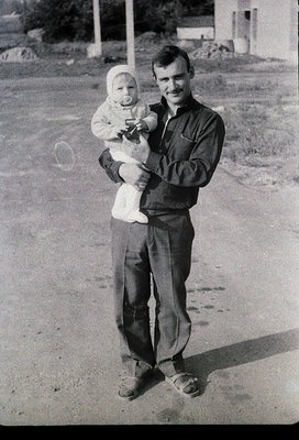 Mid-20th century black-and-white portrait of a man holding an infant outdoors, likely 1950s–1960s. Man wears button-up shirt,...