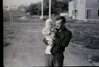 A man in mid-20th-century attire holds a baby outdoors in a residential area. The man wears a dark jacket and glasses; the ba...