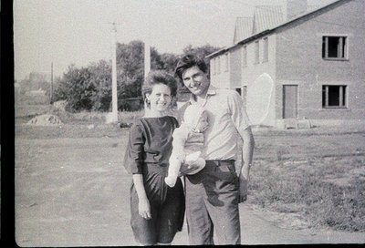 Vintage black-and-white photo of two adults posing outdoors with a child, likely mid-20th century. Woman in fitted dress, man...