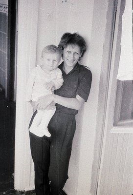 A woman in 1970s-era dress and trousers holds a toddler in a simple white shirt and diaper against a plain interior wall. War...