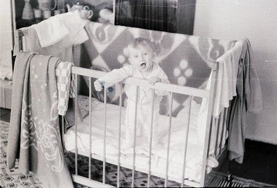 Mid-20th century black-and-white photo of a toddler in a wooden crib with vertical bars, clutching a toy. Patterned wallpaper...