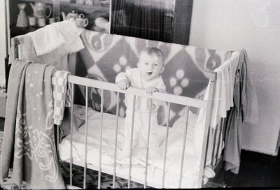 Mid-20th century black-and-white photo of a child in a classic wooden crib with floral-patterned curtains. Baby wears a simpl...