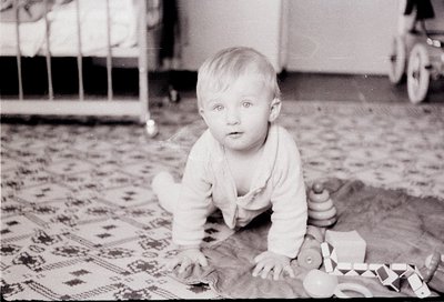Vintage black-and-white photo of a toddler crawling on patterned rug, surrounded by wooden blocks and a toy train. Mid-20th c...